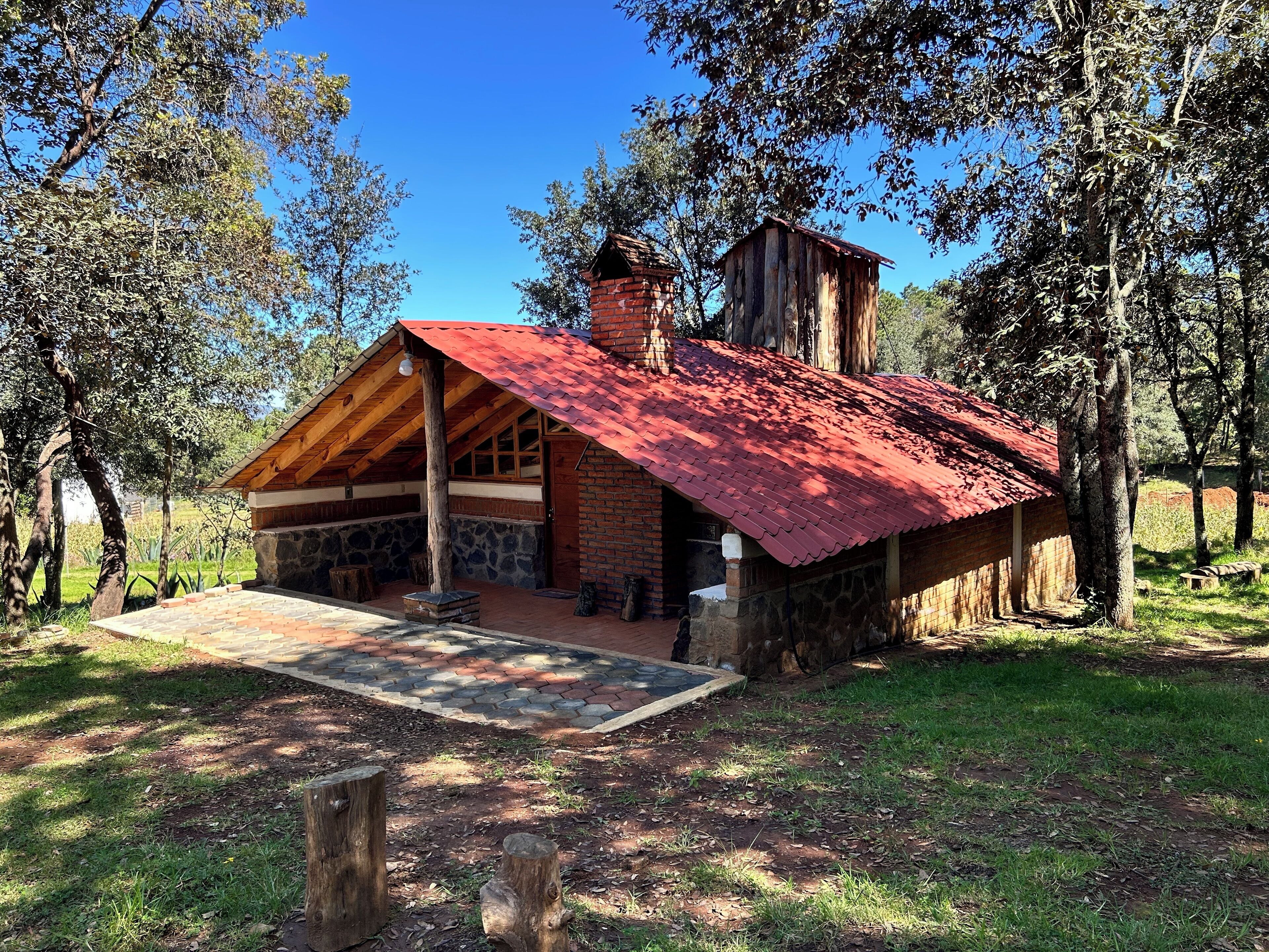 A rustic cabin with a red roof, stone walls, and a chimney. It is surrounded by trees and has a stone-paved entrance.