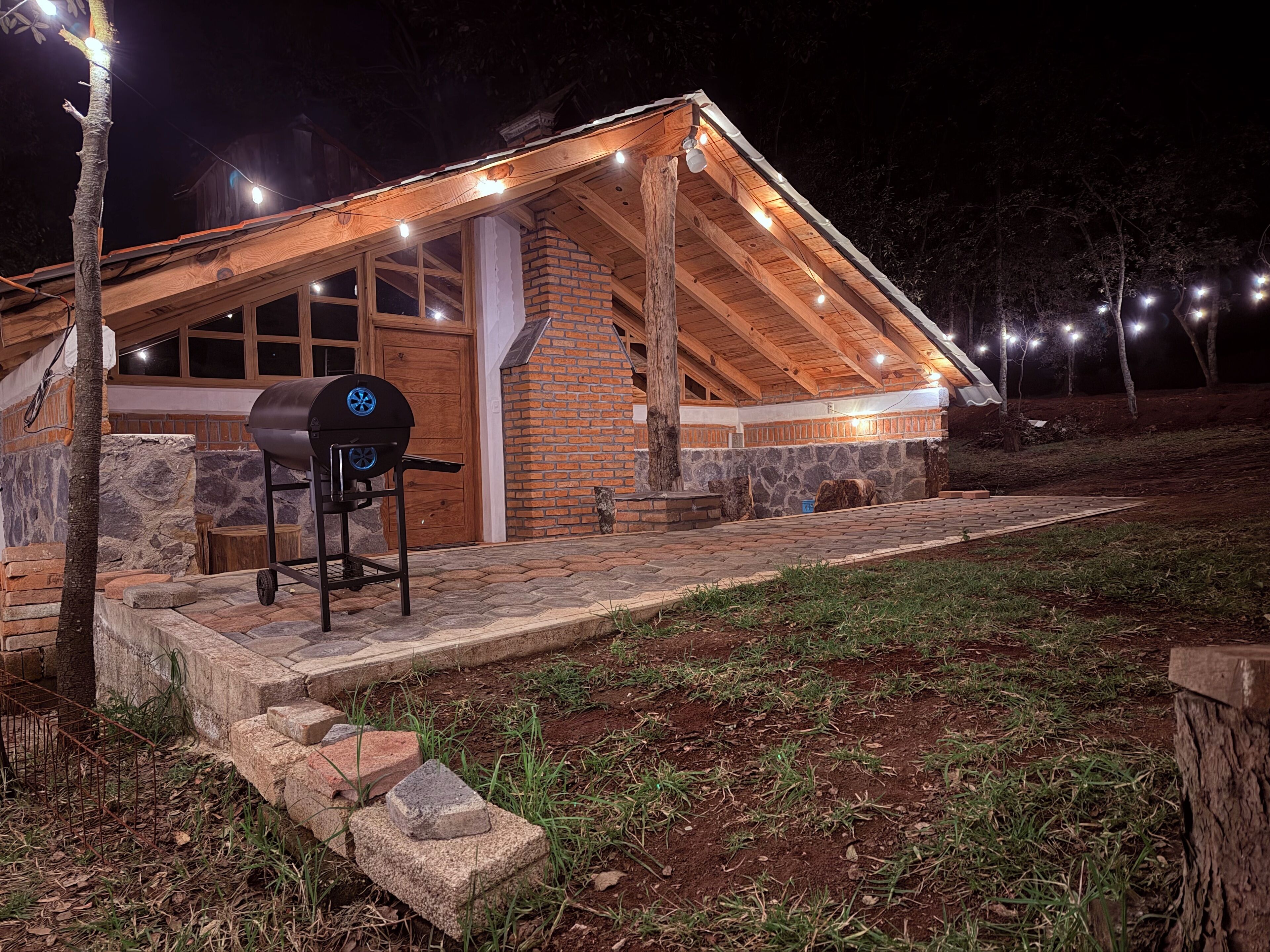 Un bâtiment rustique en bois, doté d’une cheminée en briques, d’un barbecue et d’un chemin de pierre menant à l’entrée.