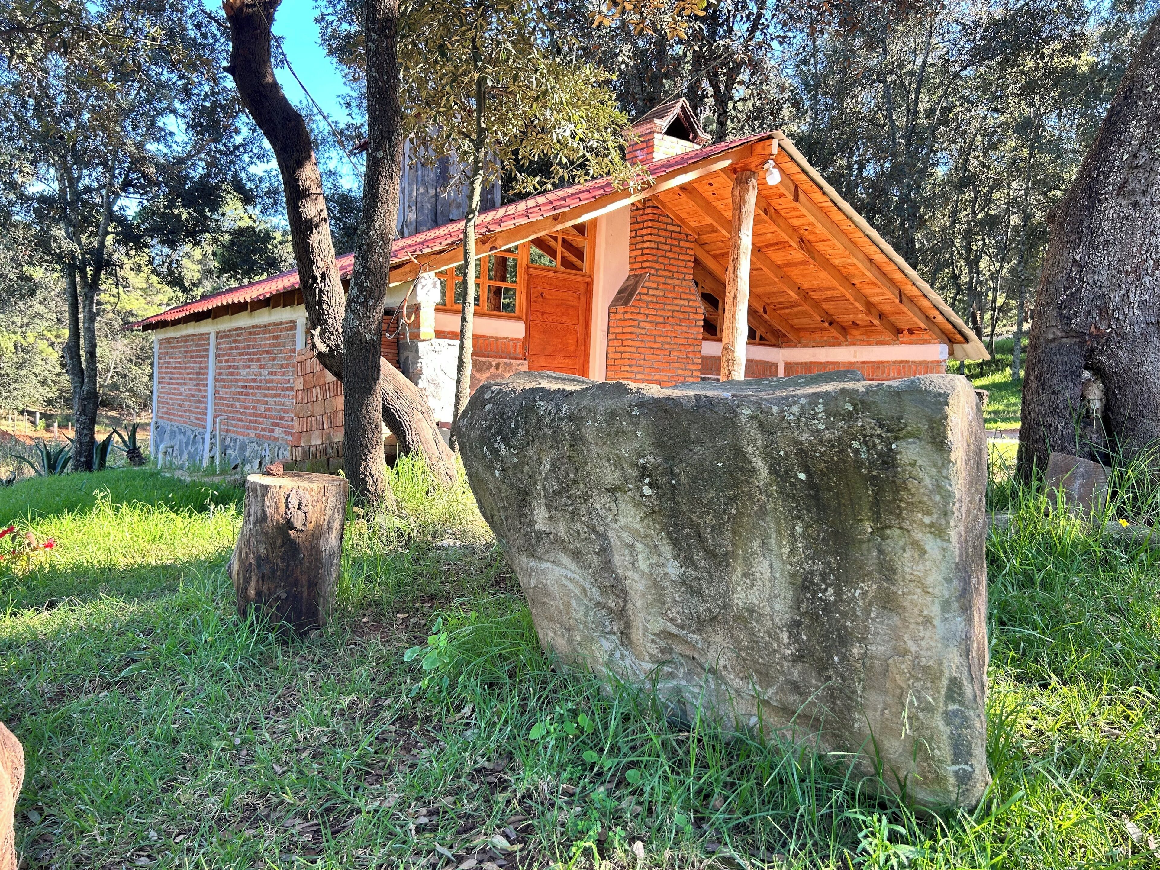 A rustic cabin with a brick exterior and wooden roof, surrounded by trees and a large rock.