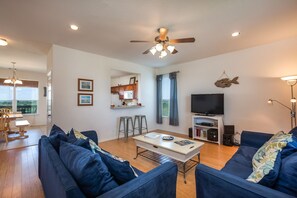 A modern living room with a sofa, coffee table, and TV. There is a kitchen area with a microwave and stools. - Sandy Hook by Ryson Vacation Rentals (Galveston)