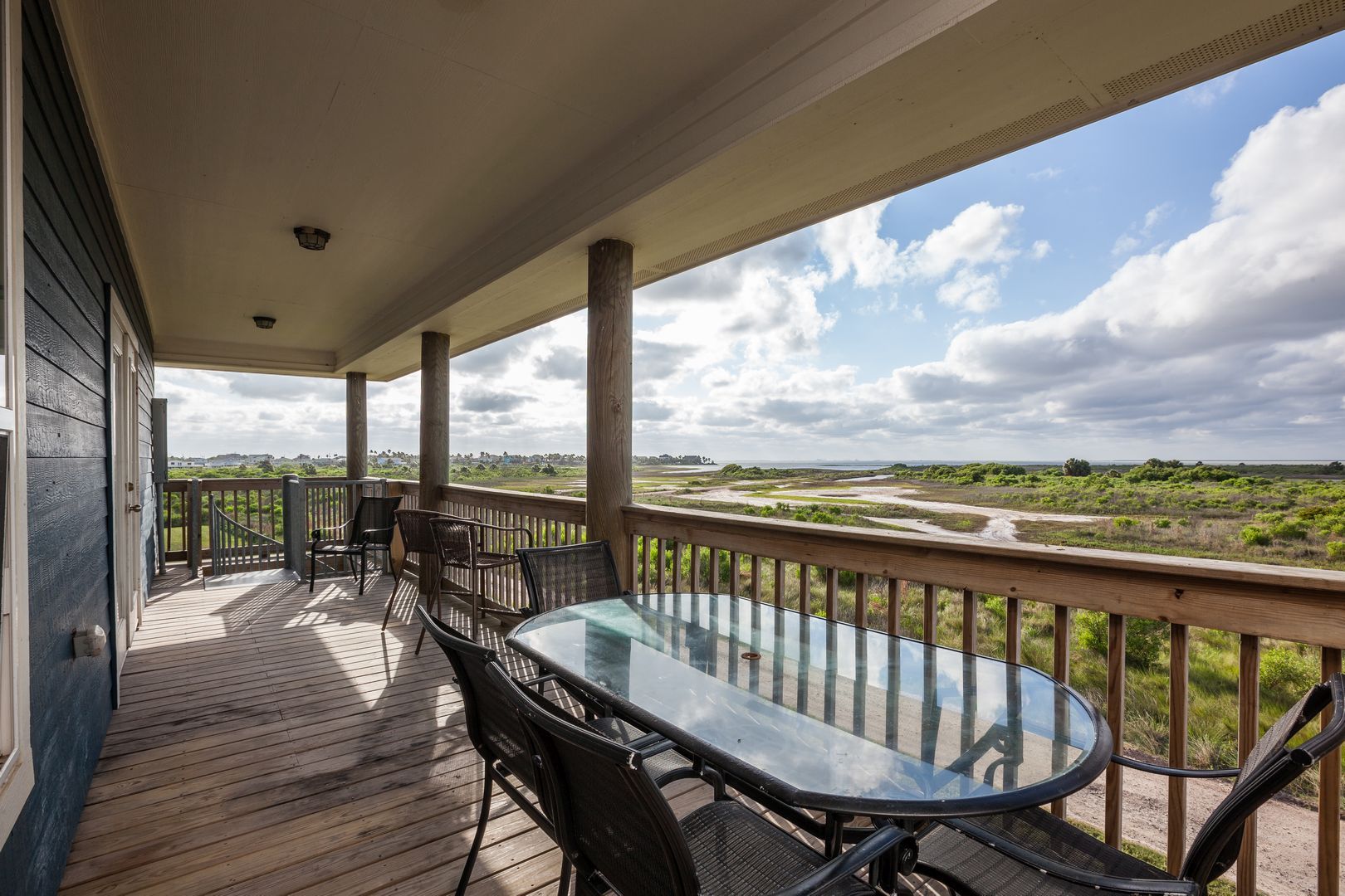 Un espace extérieur abrité, doté d’une table et de chaises en verre, d’une terrasse en bois et offrant une vue sur un paysage côtier.