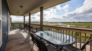 Een overdekt buitengebied met een glazen tafel en stoelen, een houten terras en uitzicht op een kustlandschap.