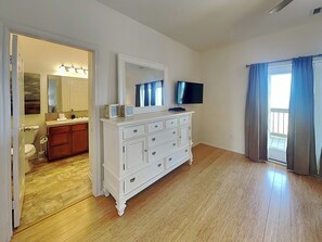 A room with a white dresser, a television, a bathroom with a sink and toilet, and a view of the outdoors through a window. - Sandy Hook by Ryson Vacation Rentals (Galveston)