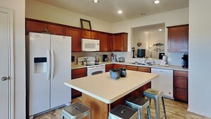 A modern kitchen with wooden cabinets, a central island, bar stools, and a refrigerator. - Sandy Hook by Ryson Vacation Rentals (Galveston)