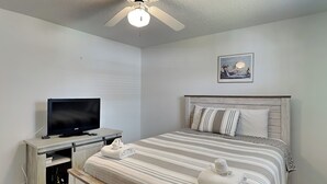 Une chambre à coucher avec un lit, un téléviseur posé sur un meuble, un ventilateur de plafond et un tableau encadré accroché au mur.