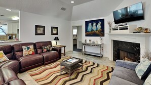 A spacious living room with a brown leather sofa, a grey armchair, and a patterned rug. A flat-screen TV is mounted above a fireplace, and there are framed pictures on the wall. - Casa de Reyes by Ryson Vacation Rentals (Galveston)
