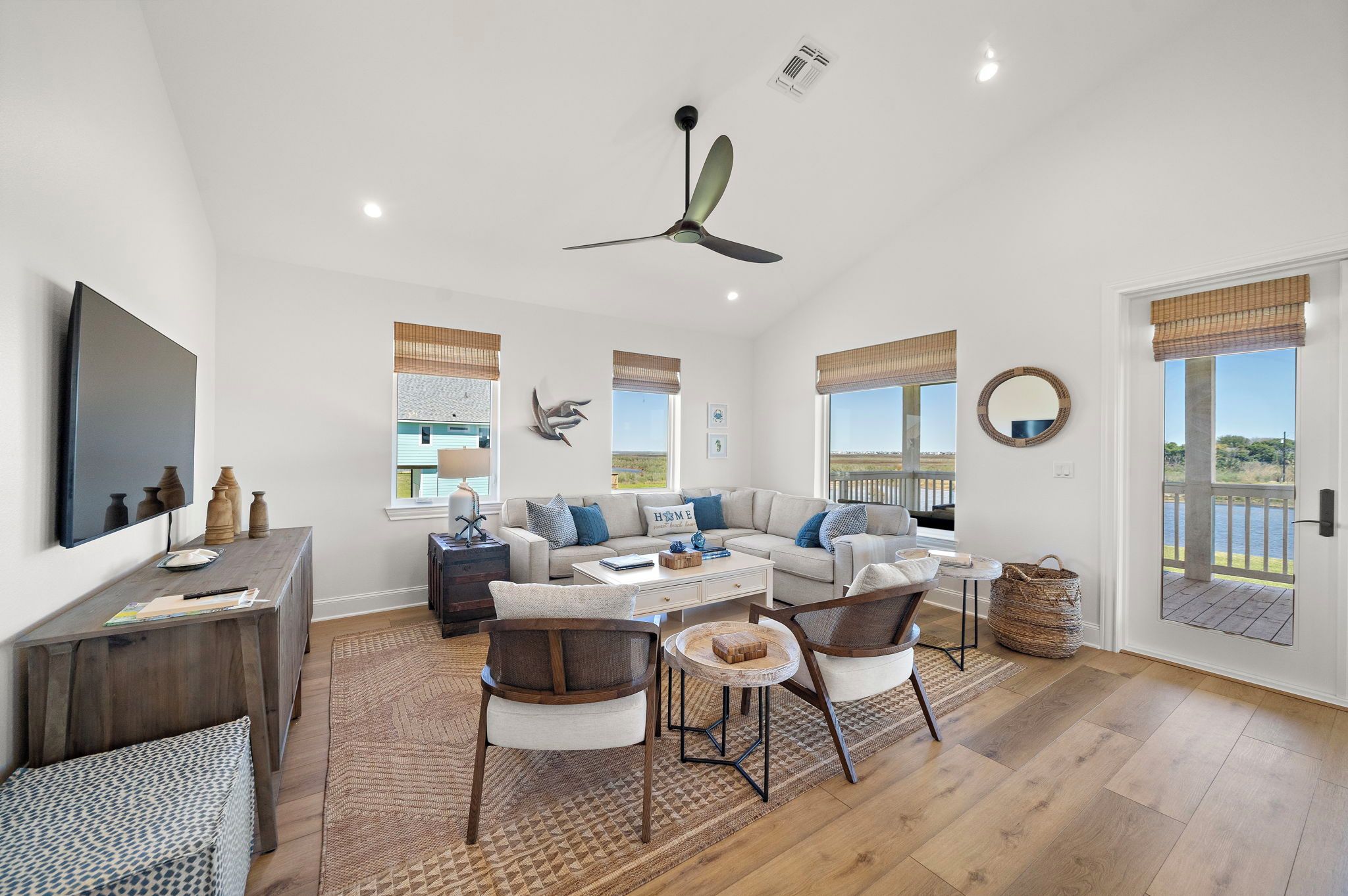 A spacious living room with a large flat-screen TV, a ceiling fan, and a view of the outdoors through large windows.
