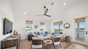 A spacious living room with a large flat-screen TV, a ceiling fan, and a view of the outdoors through large windows. - Pelican Perch by Ryson Vacation Rentals (Galveston)