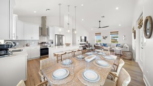 A modern kitchen with white cabinetry, stainless steel appliances, and a dining area with a wooden table set for six. - Pelican Perch by Ryson Vacation Rentals (Galveston)