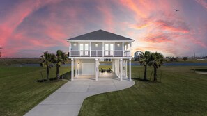 A two-story house with a balcony, a covered patio, and a view of a body of water during sunset.
