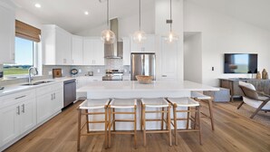 A modern kitchen with white cabinets, a large island, hanging pendant lights, and a dining area with bar stools. - Pelican Perch by Ryson Vacation Rentals (Galveston)