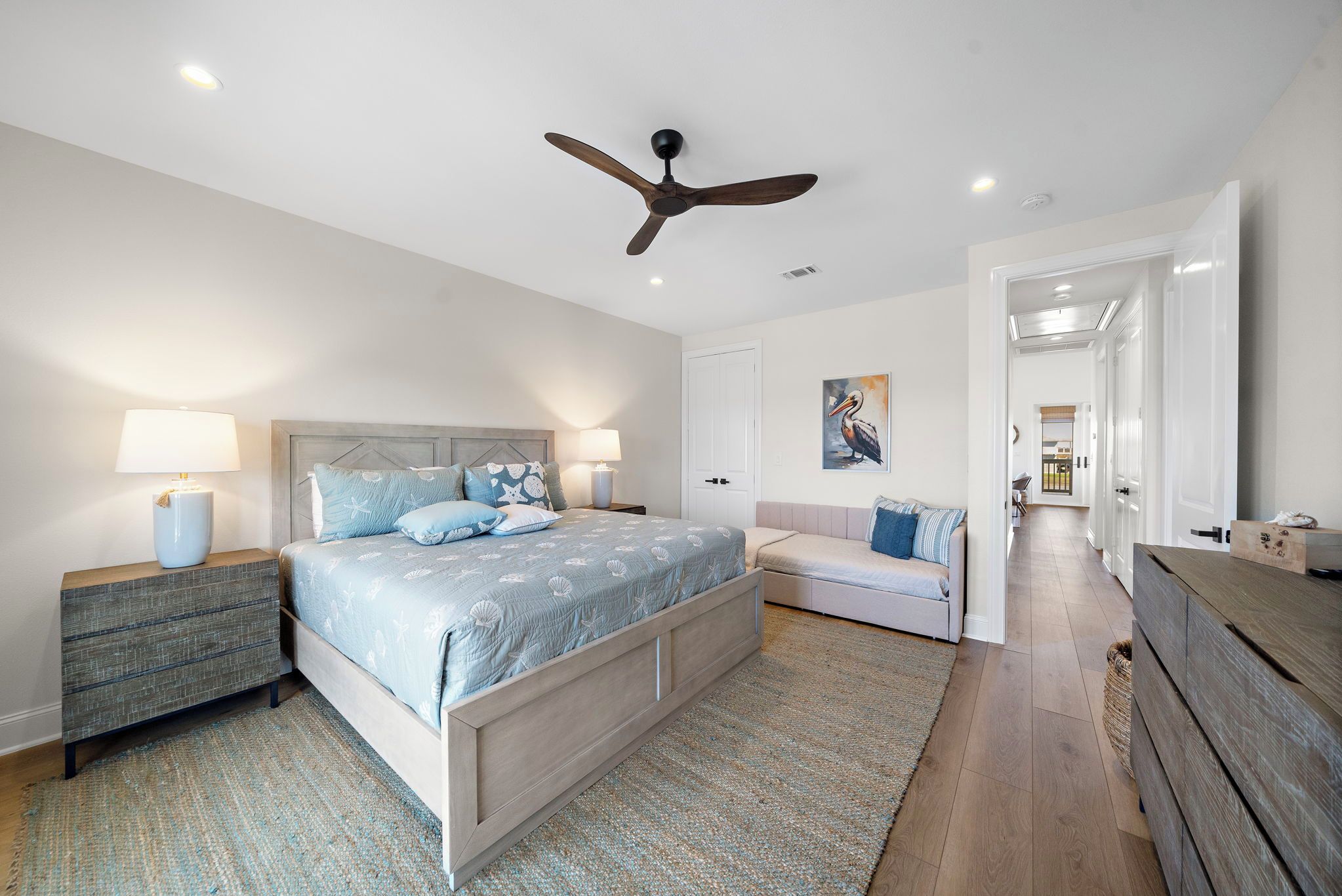 A bedroom with a bed, bedside tables, a ceiling fan, a door, and a hallway leading to another room.