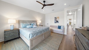 A bedroom with a bed, bedside tables, a ceiling fan, a door, and a hallway leading to another room. - Pelican Perch by Ryson Vacation Rentals (Galveston)