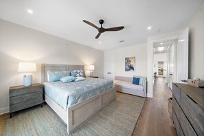 A bedroom with a bed, bedside tables, a ceiling fan, a door, and a hallway leading to another room. - Pelican Perch by Ryson Vacation Rentals (Galveston)