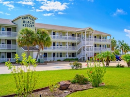 Ein mehrstöckiges Hotel mit Balkonen, einem Schild mit der Aufschrift 'Kontiki Beach Resort' und einem gepflegten Rasen mit Pflanzen und einem Felsgestalt.