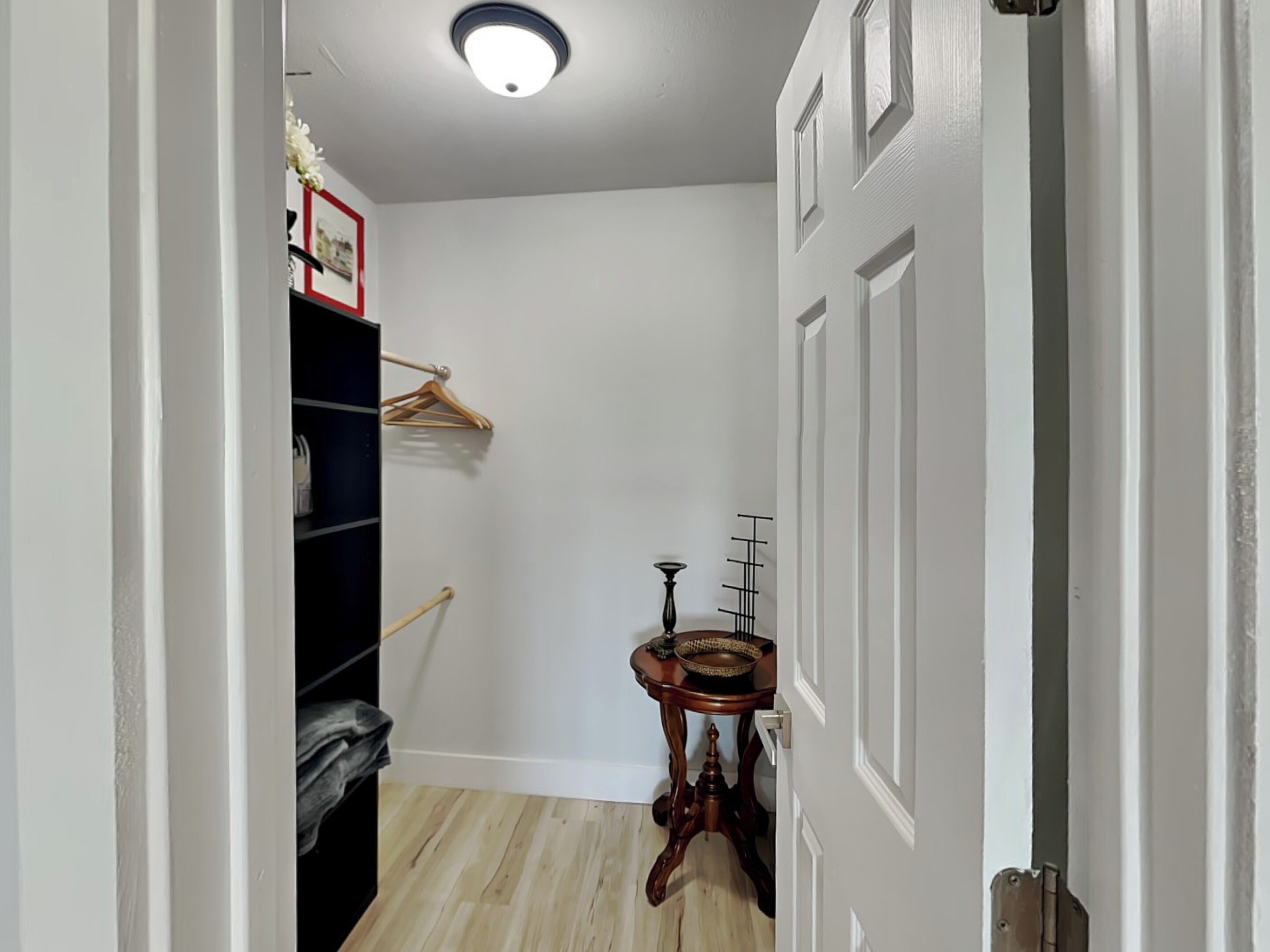 Un couloir doté d’un plancher en bois, une étagère noire contenant des vêtements, une table en bois avec un panier, et une porte surmontée d’un luminaire.