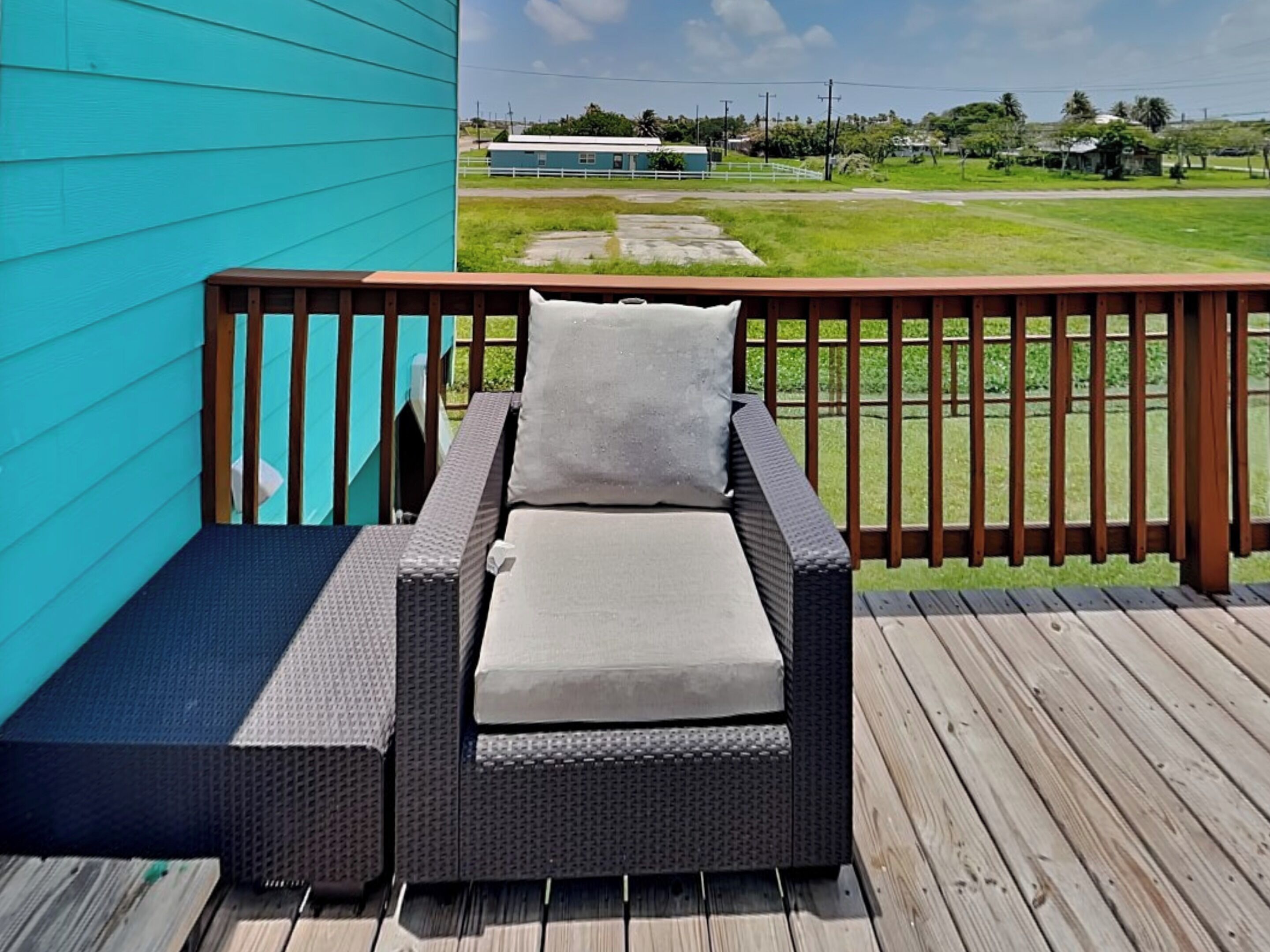 Ein Korbsessel mit Kissen auf einer Holzterrasse, mit einer blauen Wand und Blick auf eine Rasenfläche sowie Gebäude im Hintergrund.