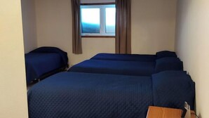 Ein Zimmer mit drei nebeneinander liegenden Betten, einem fenster mit Vorhängen und einem Nachttisch aus Holz mit einer Lampe.