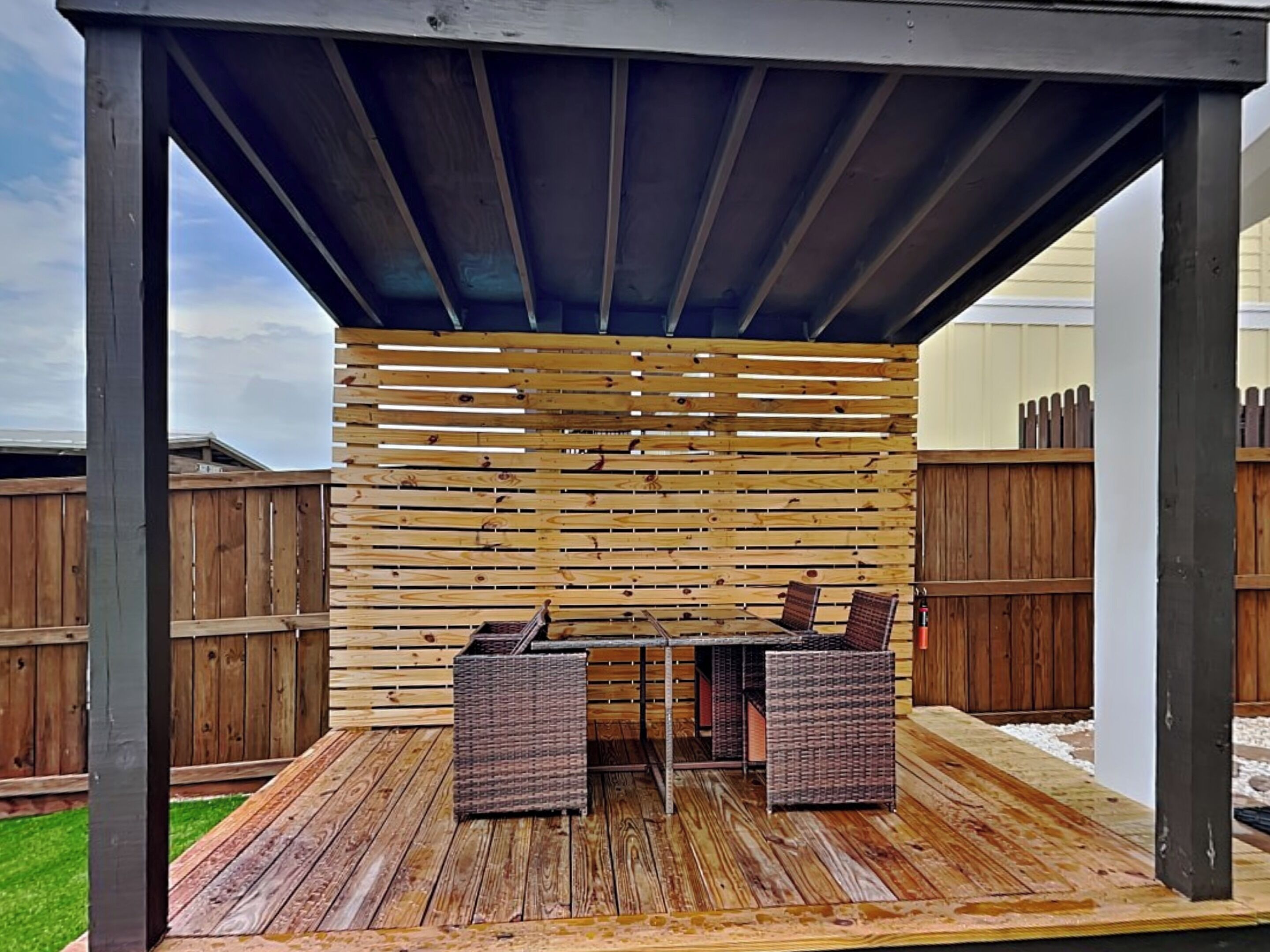 An outdoor patio with a wooden deck, a covered seating area with wicker chairs, and a wooden fence in the background.