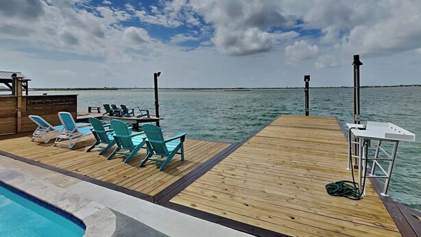 Un terrazzo in legno con poltrone relax, una piscina e vista sul mare sotto un cielo parzialmente nuvoloso.