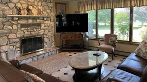 A living room with a stone fireplace, a flat-screen TV, a sofa, and a large window with a view of greenery. - Spacious Vacation 4BR Home Near Southold Beach, Greenport, and Top Wineries (Southold)