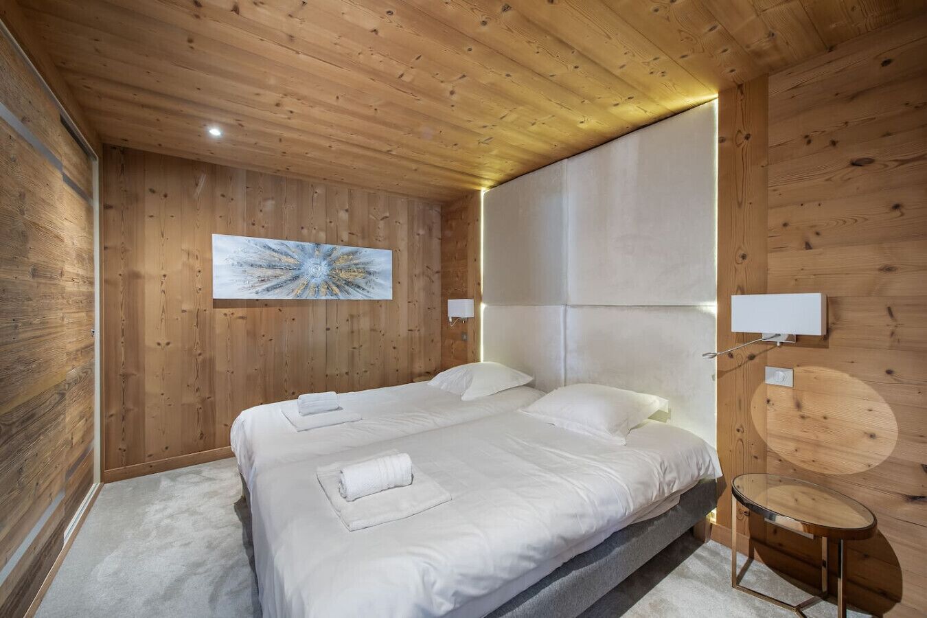 Une pièce aux murs lambrés, avec un lit, une lampe fixée au mur et une table ronde.