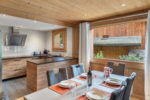 Cozinha moderna com ilha central, área de jantar com mesa posta para quatro e uma grande janela com vista para uma estrutura de madeira lá fora.
