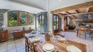 Een ruime eetruimte met een grote houten tafel, gedekt voor een maaltijd, stenen muren en grote ramen met uitzicht op groen.
