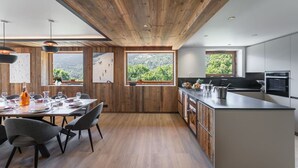 Una cocina moderna con vigas de madera en el techo, ventanas amplias con vistas a la vegetación y un comedor con mesa preparada para cuatro.