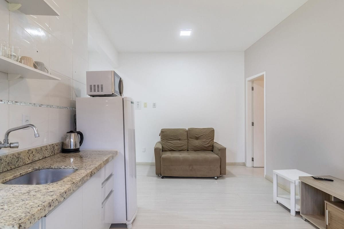A compact kitchen with a sink, microwave, and kettle, a beige sofa in a hallway, and a TV on a wooden stand.
