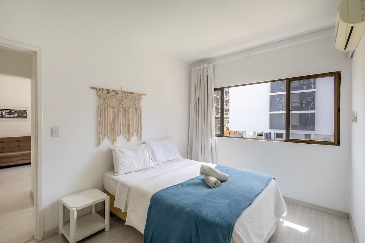 A minimalist bedroom with a bed, a small bedside table, a macrame wall hanging, and a window with curtains.