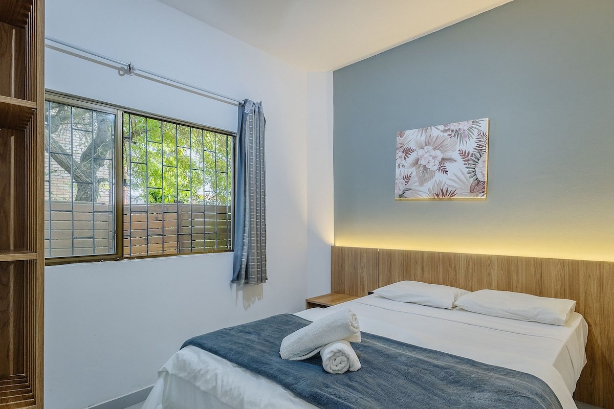 A bedroom with a bed, a window with a view of greenery, a wall-mounted artwork, and a wooden headboard.