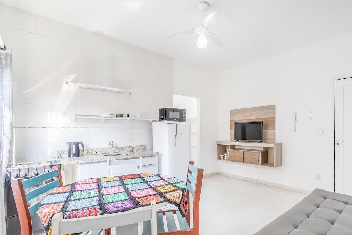 A modern kitchen with white cabinets, a microwave, and a stove. There's a dining table with a colorful tablecloth and chairs. A television is mounted on the wall, and a sofa is in the living area.