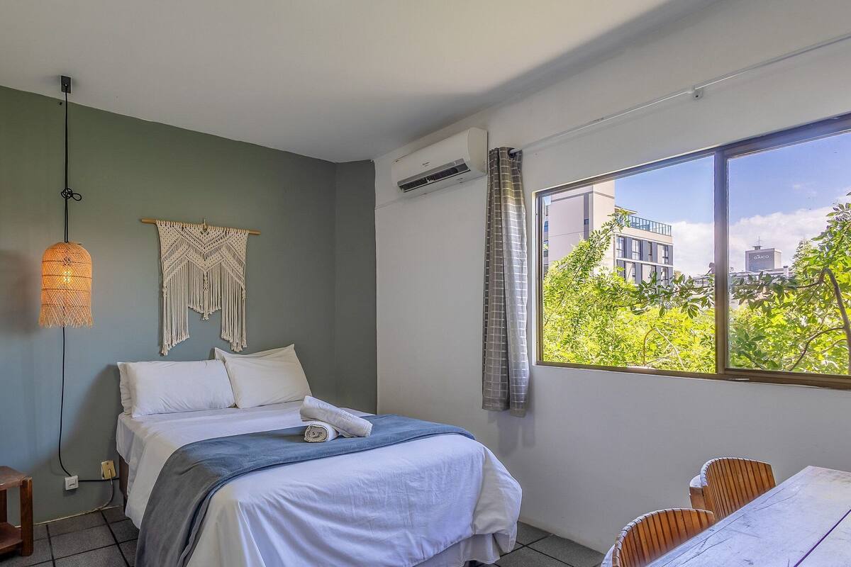 A bedroom with a bed, a desk, a chair, a hanging macrame wall decoration, and a window with a view of buildings and trees.