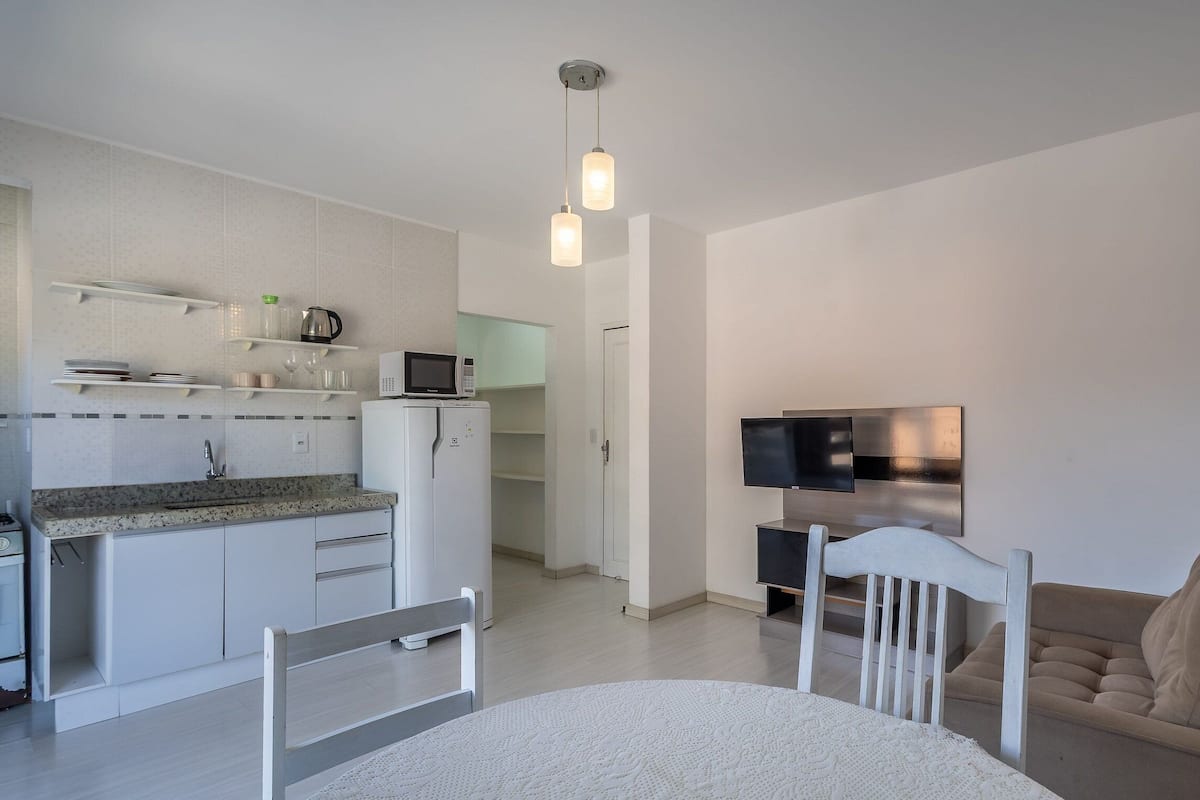 A modern kitchen with white cabinets, a microwave, and a refrigerator. There is a dining area with a white table and chairs, and a sofa in the living room.