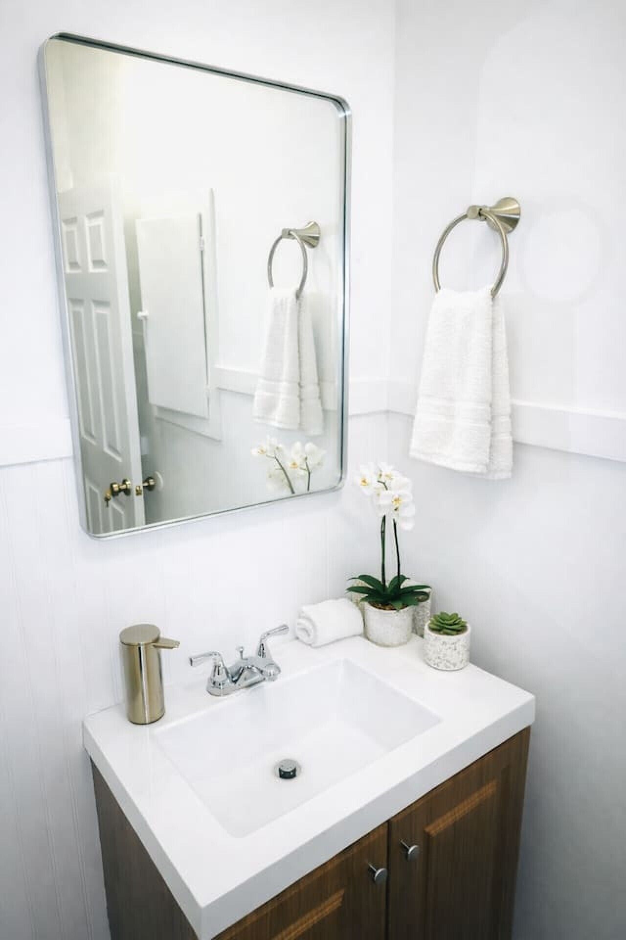 A bathroom with a white sink, a large mirror, two towels hanging on towel rings, a potted orchid, and a gold-colored pitcher.