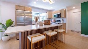 A modern kitchen with a central island, wooden bar stools, stainless steel appliances, and a unique ceiling light fixture.