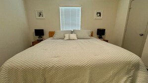 A bedroom with a large bed, two bedside tables with lamps, a window with blinds, and two framed pictures on the wall.