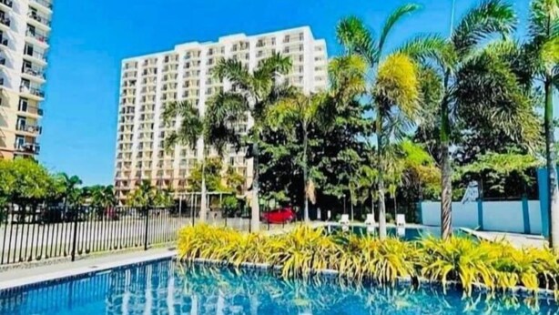 A swimming pool with a tiled edge, surrounded by palm trees and a fence, with a high-rise building in the background. - Studio with Balcony at Saekyung 956 (Lapu-Lapu City)