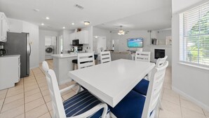 Una cocina moderna con armarios blancos, una zona de comedor con una mesa blanca y sillas con cojines azules, un horno empotrado y un televisor en la pared.