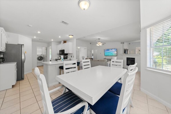 Una cocina moderna con armarios blancos, una zona de comedor con una mesa blanca y sillas con cojines azules, un horno empotrado y un televisor en la pared.