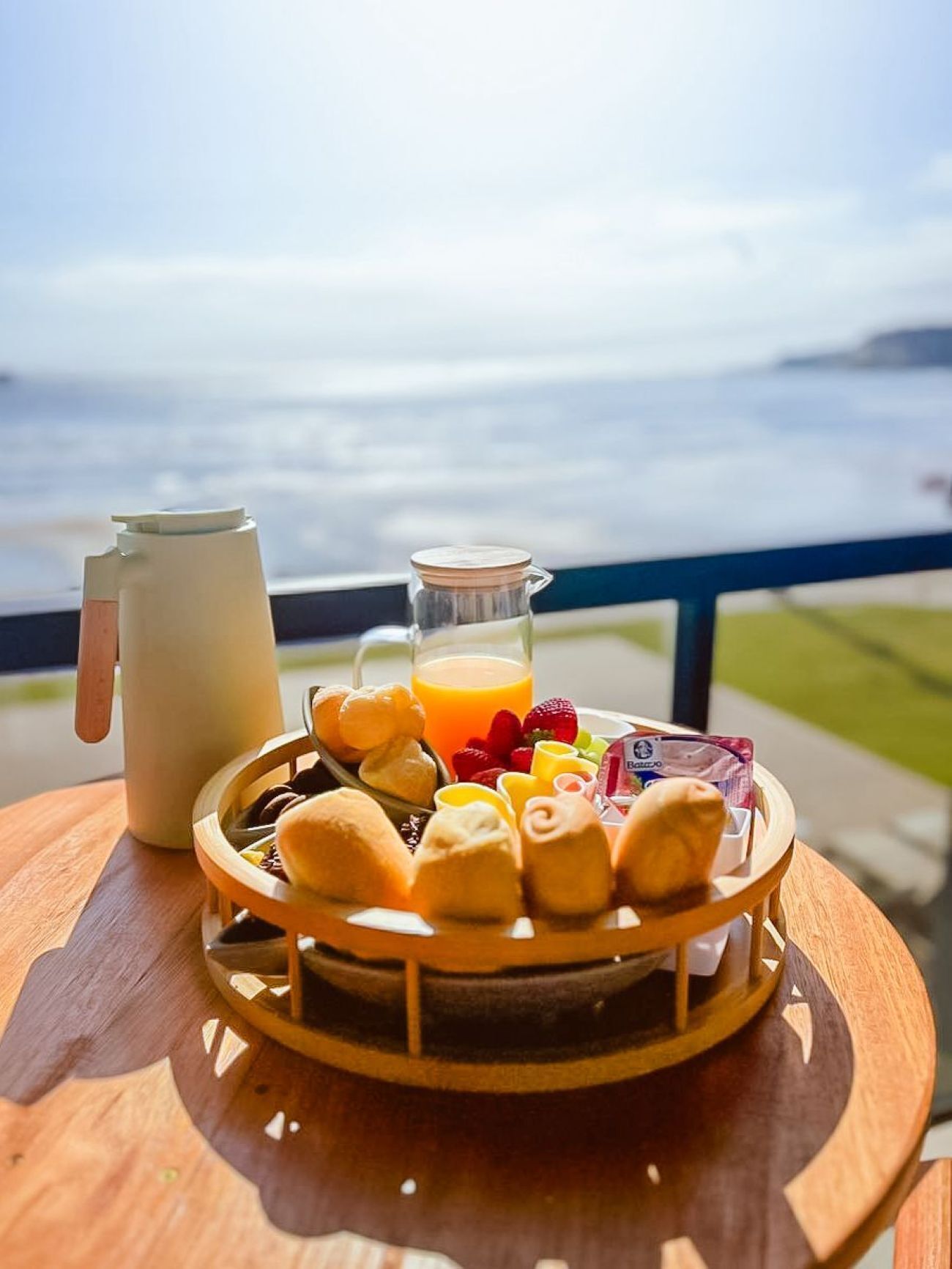 Bandeja de desayuno con pastelería, frutas y jugo sobre una mesa de madera con vista al mar.