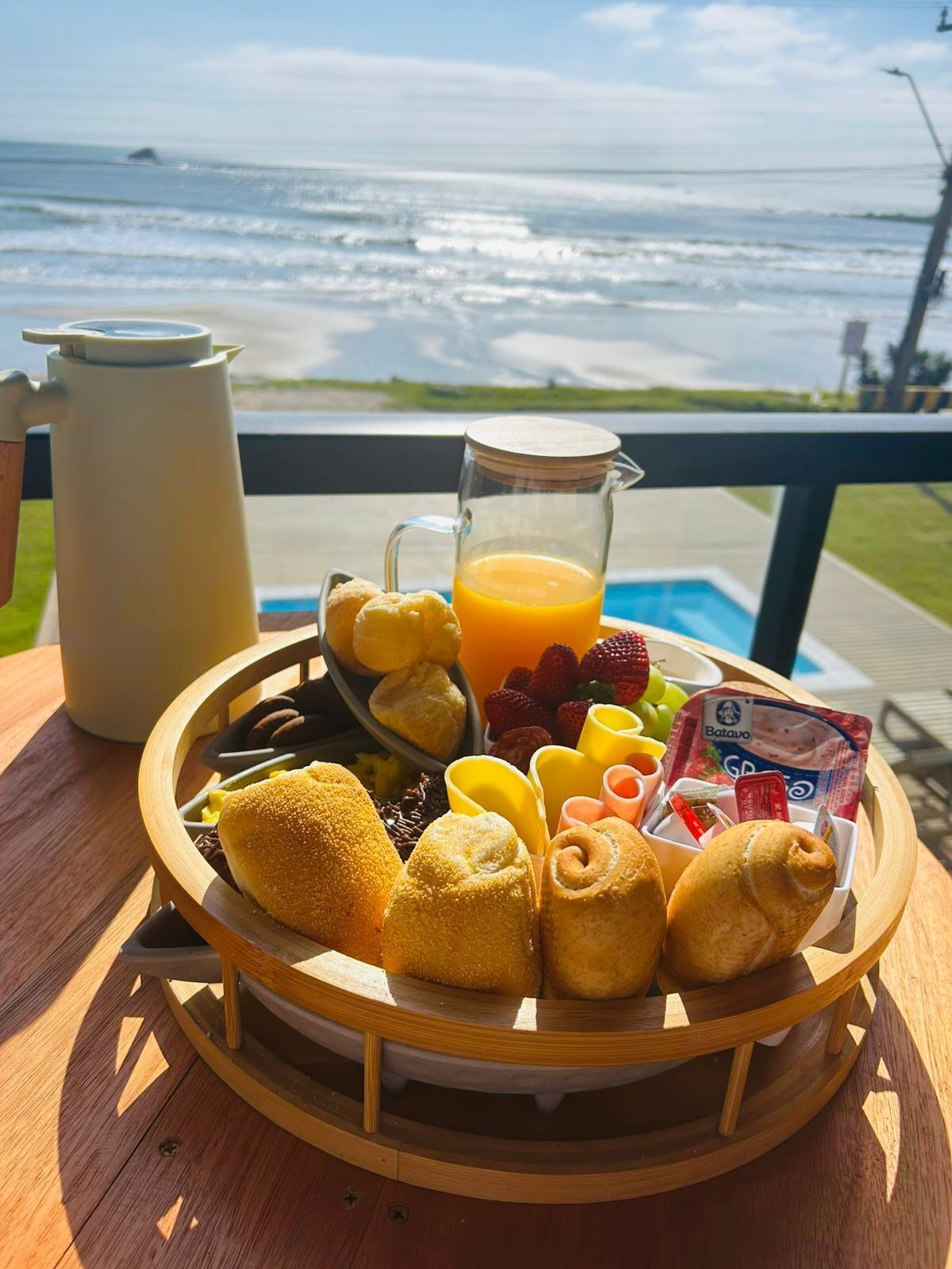 Bandeja de desayuno con pasteles, frutas y jugo sobre una mesa de madera junto a una piscina con vista al océano.