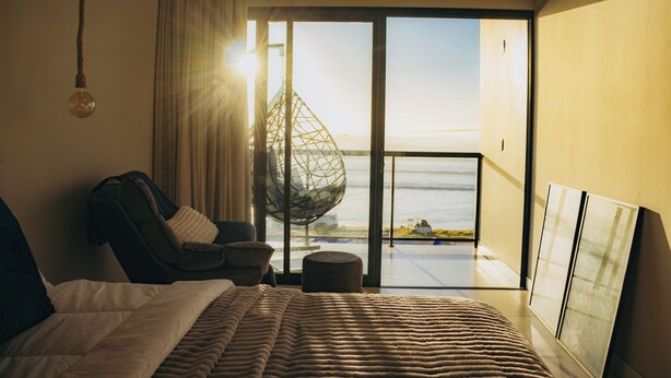 A modern hotel room with a large bed, a seating area with a chair and ottoman, a hanging light fixture, a large window with a view of the sea, and a glass door leading to a balcony. - Pousada Austral Suítes (Imbituba)