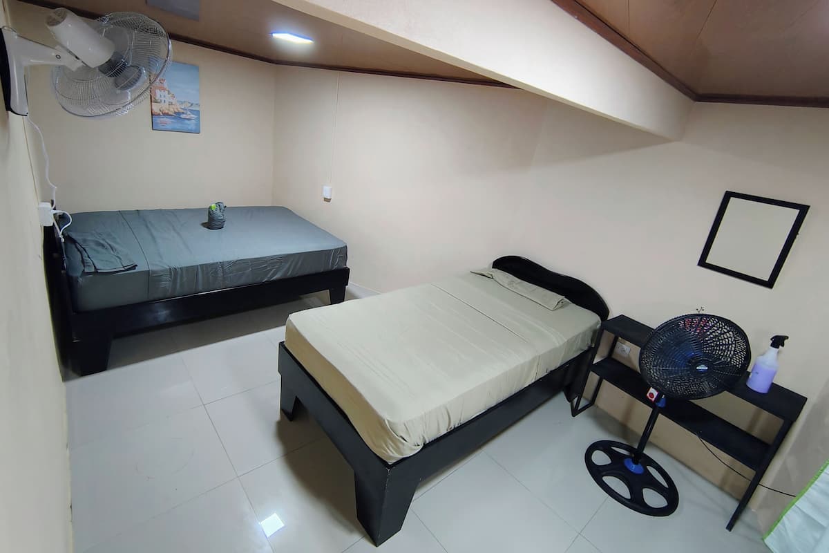 A small, well-lit room with two beds, a ceiling fan, and a wall-mounted picture.