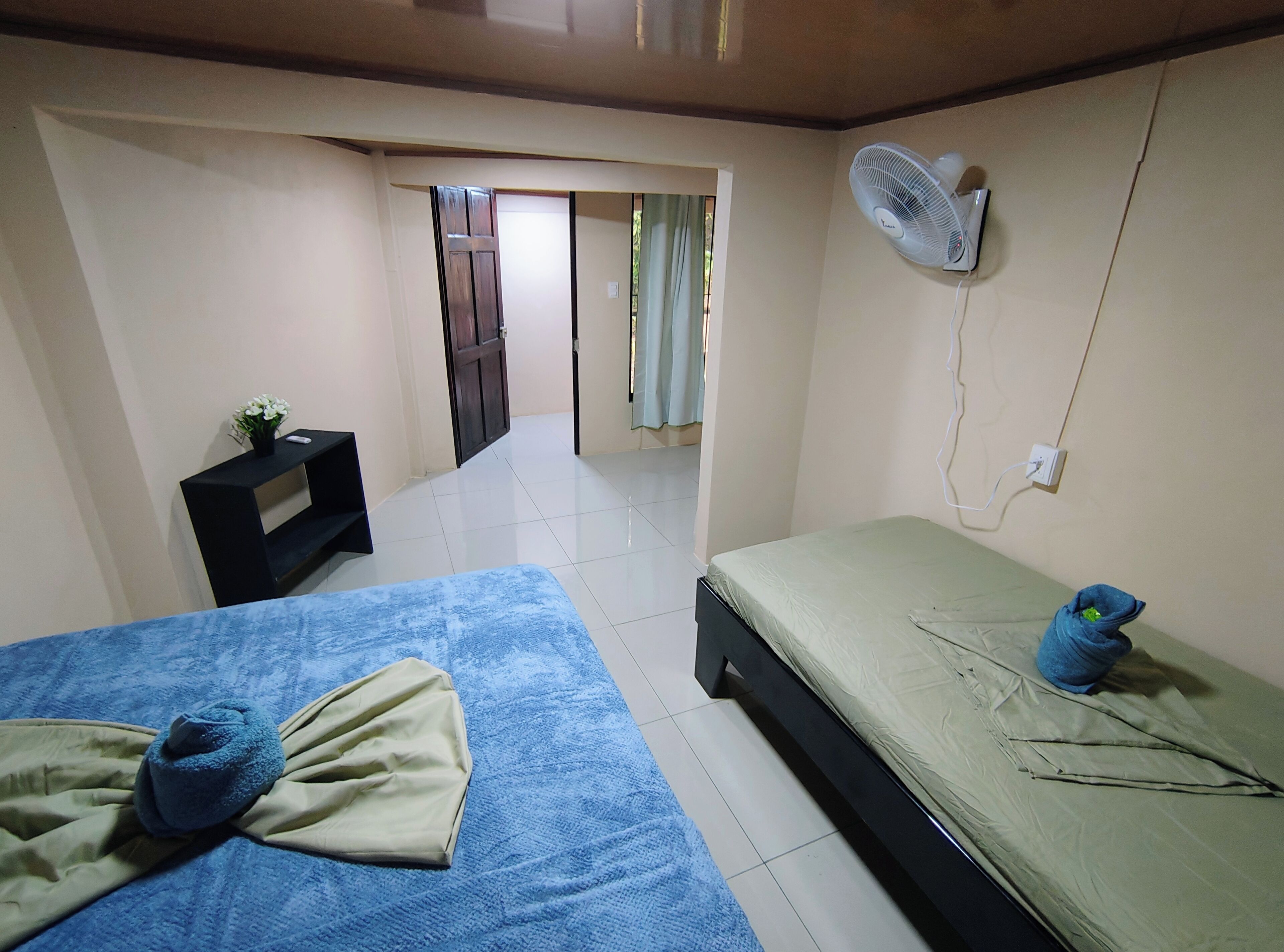 A room with two beds, a bedside table, a wall-mounted fan, and a door leading to another room.