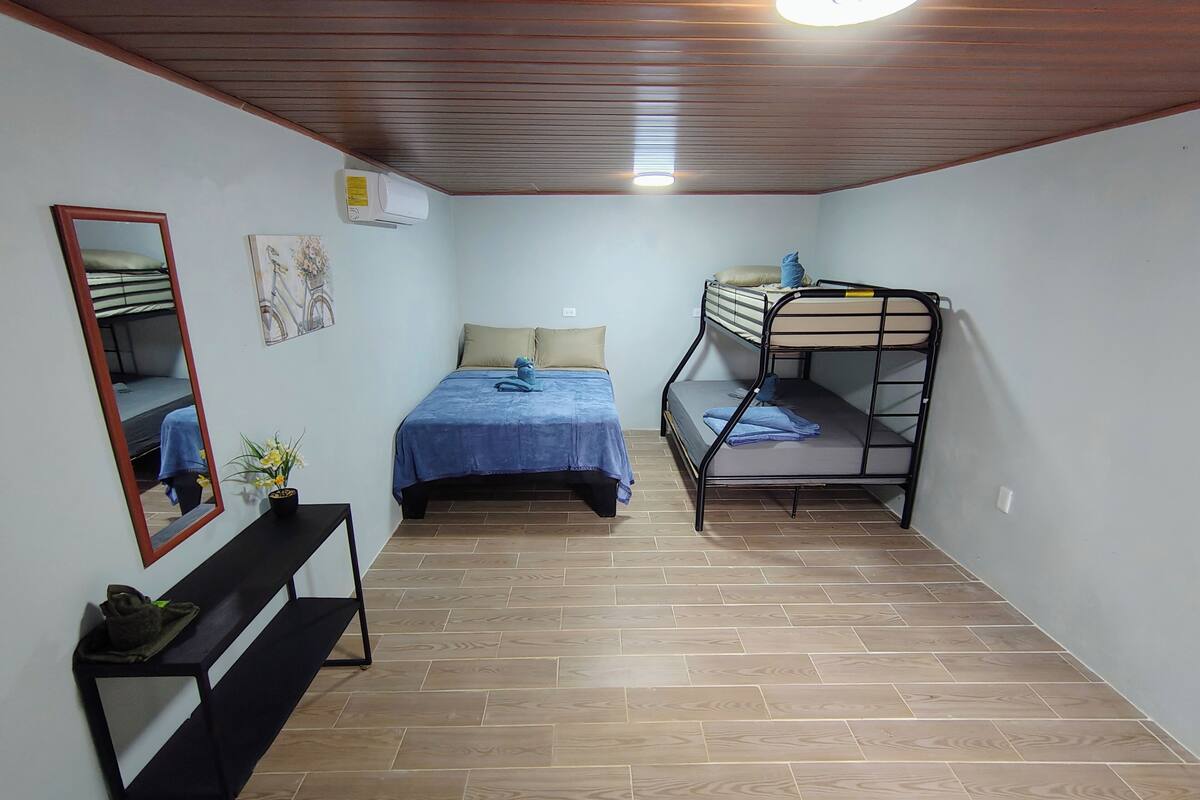 A small, well-lit room with a bunk bed, a single bed, a mirror, a small table with a plant, and a ceiling fan.
