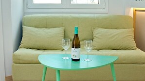 A room with a light green sofa, a small turquoise table with a bottle of wine and two glasses, and a window with a view of buildings. - Emily in Athens - Central Veranda flat (Athina)