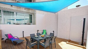 An outdoor patio with a table set for four, surrounded by chairs, a sun lounger, and a blue shade sail. - Emily in Athens - Central Veranda flat (Athina)