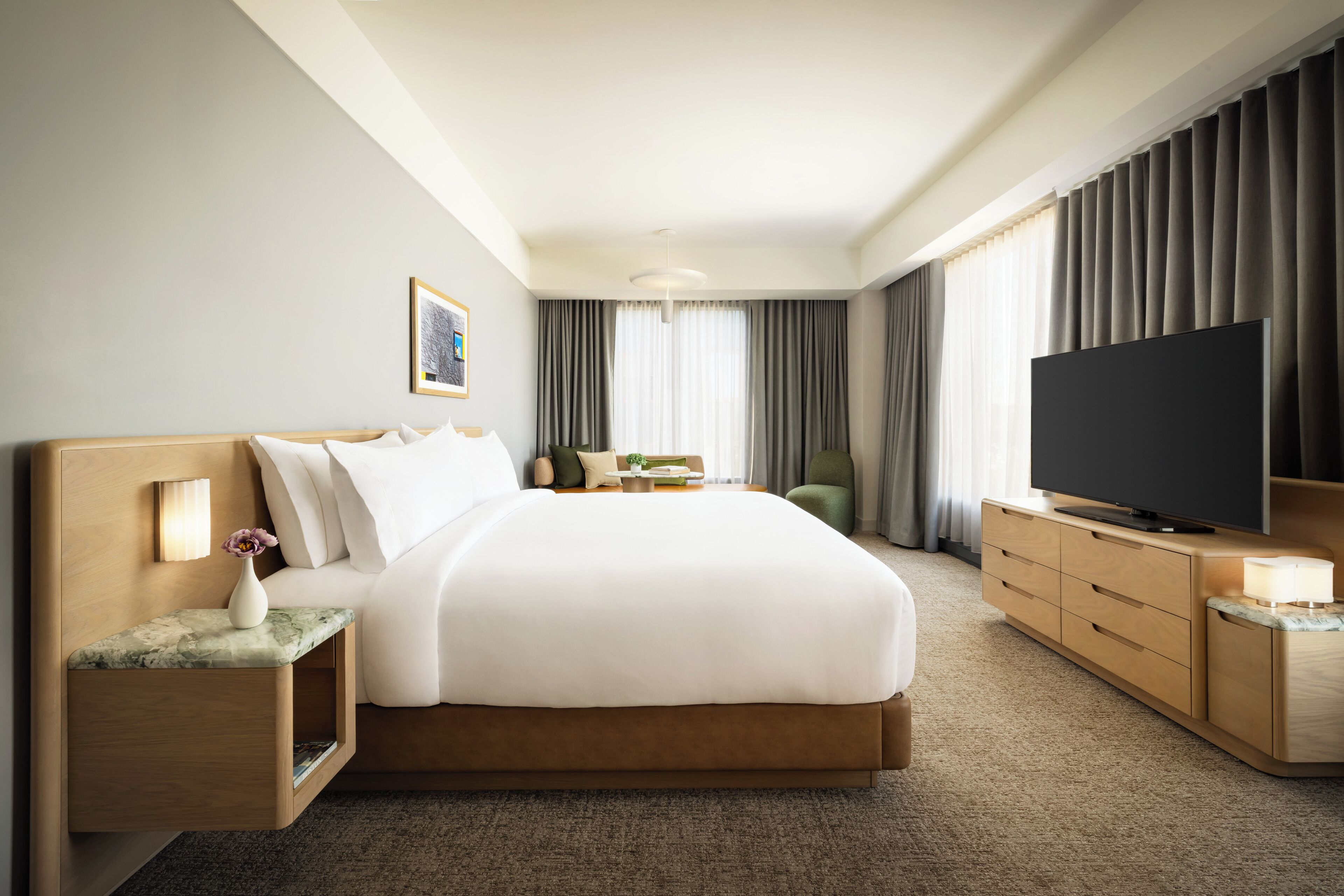 A hotel room with a large bed, a flat-screen TV, a bedside table with a lamp, a small side table with a vase, and a window with curtains.